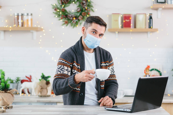 Sick man with a cold in a protective mask working remotely from home using a laptop during the New Year and Christmas holidays - Fotografie, Obrázek