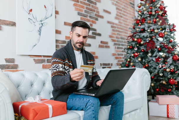 A man with a beard sitting on the couch during the New Year holidays, nervous and worried trying to get a loan online, using a laptop and credit card - Valokuva, kuva