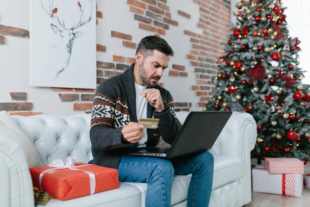 A man with a beard sitting on the couch during the New Year holidays, nervous and worried trying to get a loan online, using a laptop and credit card - Zdjęcie, obraz