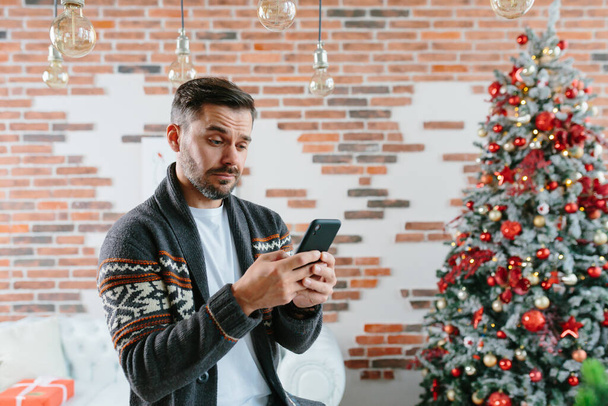 A serious and focused man is corresponding on a mobile phone near the Christmas tree at home - Photo, image