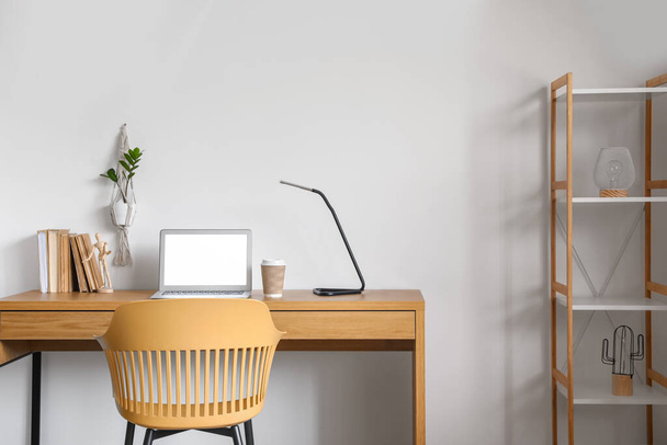 Робоче місце з сучасним ноутбуком, лампою та паперовою чашкою кави на дерев'яному столі біля білої стіни
 - Фото, зображення