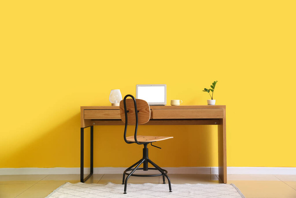 Työpaikka moderni kannettava tietokone, lamppu, kuppi ja huonekasvi lähellä keltaista seinää - Valokuva, kuva