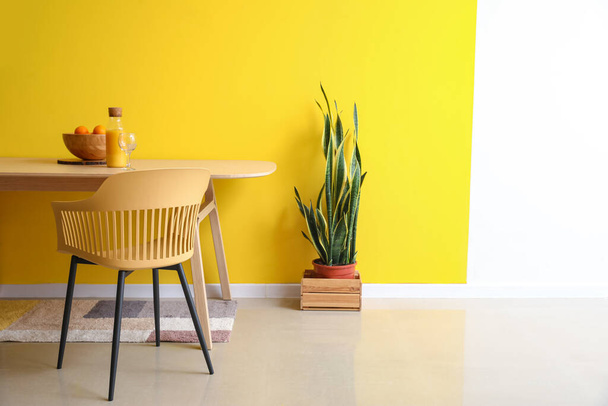 Mesa de madera con frutas y jugo, silla y planta de interior cerca de la pared de color en el comedor - Foto, imagen