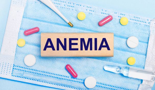 On a light blue disposable face mask there are tablets, a thermometer, an ampoule and a wooden block with the text ANEMIA. Medical concept - Foto, Imagem