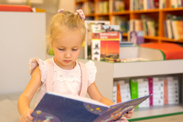 Uma menina bonita em rosa com um livro nas mãos na biblioteca não olha para a moldura - Foto, Imagem