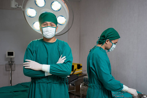 Οι γιατροί στο χειρουργείο φορώντας πράσινη μάσκα χειρουργικό φόρεμα καπέλο τρίχας λατέξ γάντι ένα περίπτερο με τα χέρια σταυρωμένα μπροστά από το κρεβάτι και ηλεκτρικό απινιδωτή άλλο, ενώ ο γιατρός προετοιμασία όργανα - Φωτογραφία, εικόνα