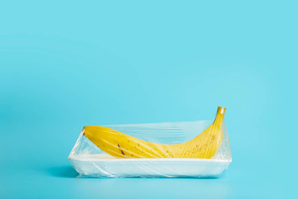 Fruits in plastic packaging from the supermarket are minimal. Banana in cellophane and non-degradable plastic on a blue background. Biodegradable product packaging, environmental protection, nature - Photo, Image