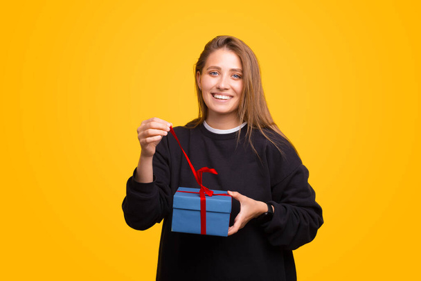 Cheerful young woman is unwrapping the gift she has received. She is standing over yellow background. - Photo, Image