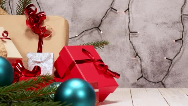 Concepto de Navidad. Saco de yute con muchos regalos de Navidad. Paquetes rojos, marrones y plateados con cintas rojas en una tabla de madera blanca. Decoraciones navideñas alrededor y luces brillantes en el fondo - Metraje, vídeo