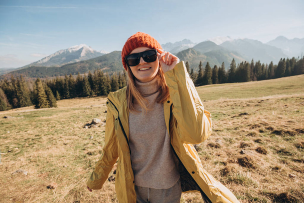 Μια νεαρή ξανθιά με γυαλιά ηλίου που χορεύει στα βουνά. Ελευθερία, ευτυχία, ταξίδια και διακοπές έννοια, υπαίθριες δραστηριότητες. Ευτυχισμένο κορίτσι με κόκκινο καπέλο και κίτρινο μπουφάν. - Φωτογραφία, εικόνα