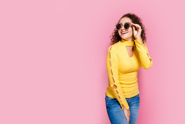beauty curly girl smiling in yellow outfit and sunglasses while standing on pink studio background - Foto, imagen