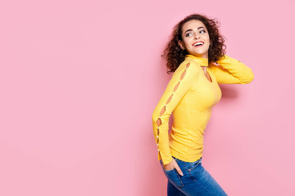 beauty smiling curly girl in yellow outfit while raises up her hand over her head on pink background - Photo, image