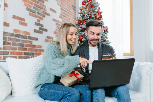 Młoda rodzina mąż i żona wybrać razem prezenty świąteczne w sklepie internetowym, siedzi w domu w pobliżu choinki z laptopem - Zdjęcie, obraz
