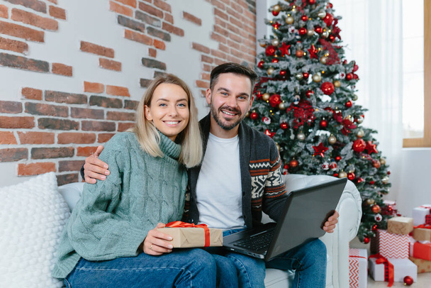 Young family husband and wife choose together Christmas gifts in an online store, sitting at home near the Christmas tree with a laptop - Fotografie, Obrázek