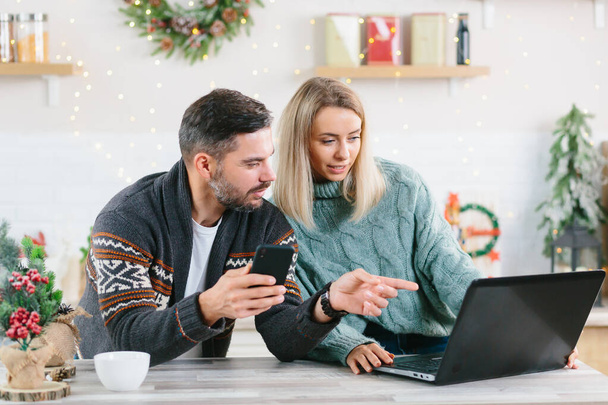 Young couple husband and wife working together online at home, with laptop sitting in the kitchen, happy together, during the New Year and Christmas holidays - Foto, afbeelding