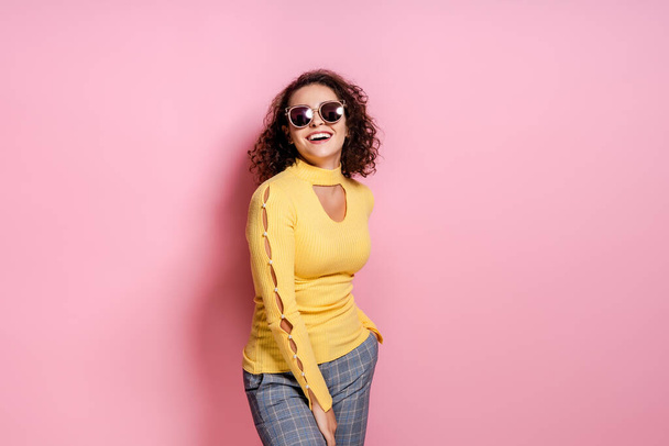 Laughing curly girl dressed yellow outfit and sunglasses while posing isolated over pink background. - Zdjęcie, obraz