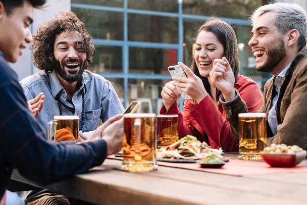 Ομάδα χαρούμενων φίλων που απολαμβάνουν μια μπύρα και φαγητό σε υπαίθρια παμπ μπαρ το απόγευμα μετά τη δουλειά, μοιράζονται καλές στιγμές στα μέσα κοινωνικής δικτύωσης με έξυπνα τηλέφωνα - Φωτογραφία, εικόνα