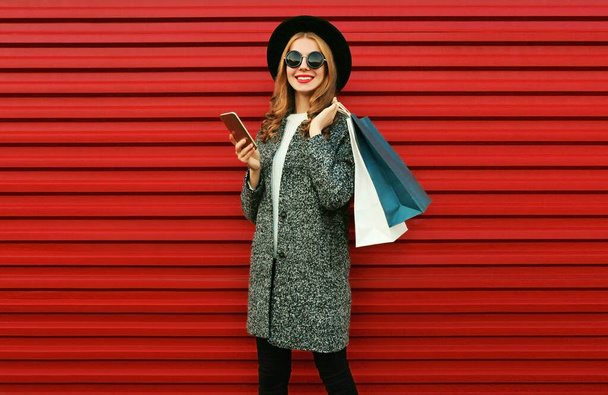 Portrait of happy smiling woman with shopping bags and smartphone wearing a gray coat, hat on colorful red background - Foto, Imagen