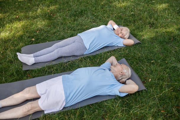 Lovely senior family couple looks at each other lying on mats after training in green park - Valokuva, kuva