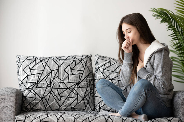 Νεαρή γυναίκα αρρωσταίνει και βήχει ενώ κάθεται στον καναπέ στο σπίτι - Φωτογραφία, εικόνα