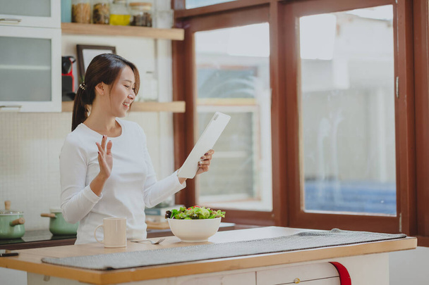 mooie aziatische vrouw groeten haar vrienden via digitale tablet in keuken voor het eten van salade groenten - Foto, afbeelding