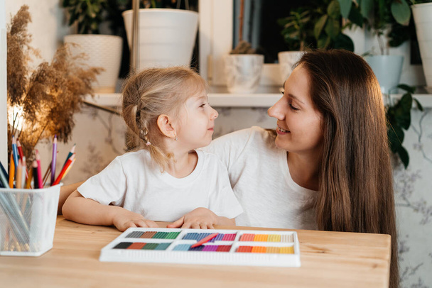 madre e hija están sentadas en una mesa con plastilina de colores para modelar, mirándose y sonriendo.El concepto del Día de la Madre y el Día del Niño. - Foto, imagen