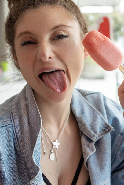 hermosa mujer que se divierte haciendo muecas y la lengua mientras come helado, detalles de la cara, estilo de vida y alegría, el uso de collar y maquillaje - Foto, imagen