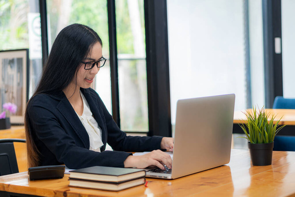 Mulher séria sentada em uma mesa e trabalhando em laptop de casa. Sorrindo mulher asiática trabalhando, lendo papéis, relatório sobre projeto freelancer feminino bem sucedido. usar um computador - Foto, Imagem