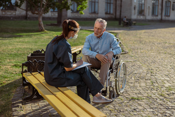Mluvící zotavující se mužský pacient na invalidním vozíku vede konverzaci se zdravotní sestrou v ochranném štítu v parku poblíž rehabilitační kliniky - Fotografie, Obrázek