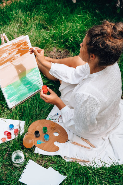 Artist painting on the easel outdoors in the garden. Open air outdoor art workshop. Draw on the canvas with brush and palette sitting on the grass during a picnic in a park - Photo, Image