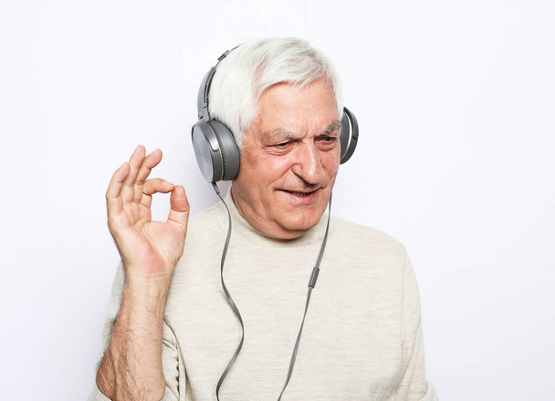 Happy old man in headphones listening to music on white background. Attractive guy in stylish sweater smiles and show ok sign. - Фото, зображення