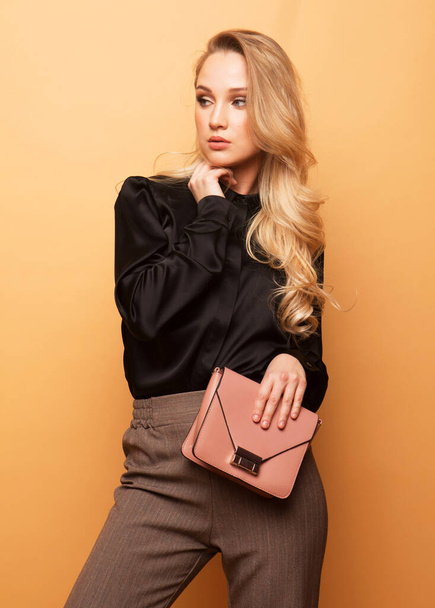 Young beautiful blond woman in a black blouse and brown pants holds a pink handbag and posing on a beige background. Fashion style. - Фото, изображение