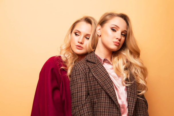 Lifestyle, fashion and people concept: Positive fashion portrait of two women, best friends posing indoor on beige background wearing winter stylish coat. Fashionable clothes. Sisters walking. - Foto, Imagen