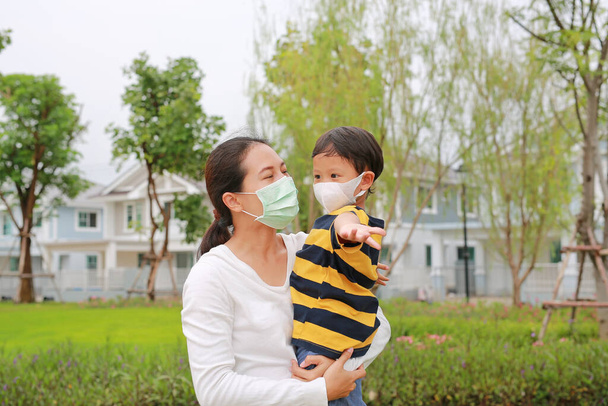 Asian mother carrying her son with wear protective face mask both in public garden during coronavirus and flu outbreak. - 写真・画像