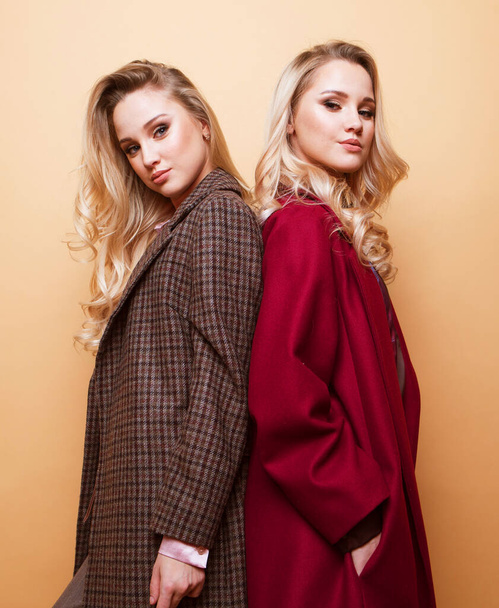 Lifestyle, fashion and people concept: Positive fashion portrait of two women, best friends posing indoor on beige background wearing winter stylish coat. Fashionable clothes. Sisters walking. - Fotó, kép