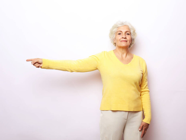 Old blonde lady smiling finger pointing on white grey background, copy space - Foto, afbeelding
