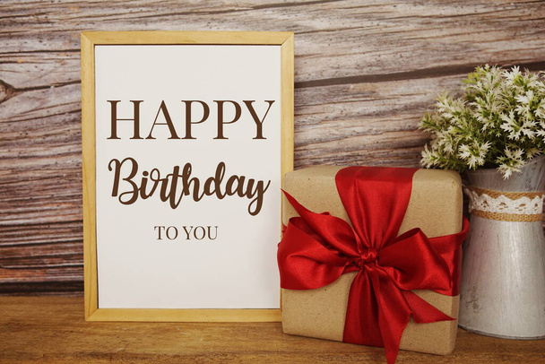 Blackboard with the text HAPPY BIRTHDAY TO YOU with gift box and red ribbon on wooden background - Fotó, kép