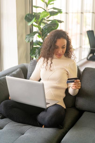 Latin woman using laptop and hand holding credit card for shopping on sofa at home - Foto, Imagem