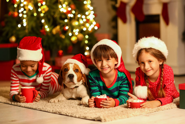 Μικρά παιδιά με φλιτζάνια ζεστό κακάο και χαριτωμένο σκυλί στο σπίτι την παραμονή των Χριστουγέννων - Φωτογραφία, εικόνα