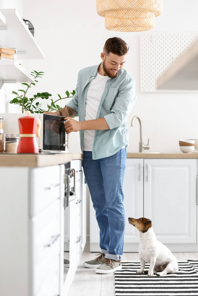 Young man with cute dog adjusting microwave oven in kitchen - Fotó, kép