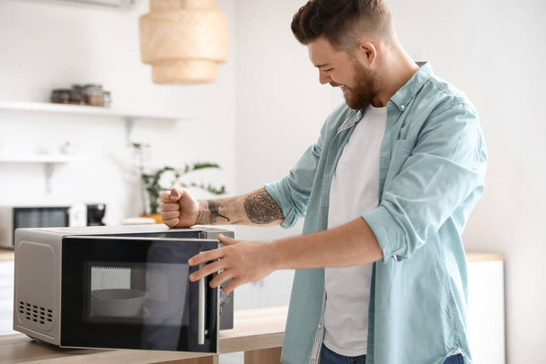 Angry young man near microwave oven in kitchen - Photo, Image