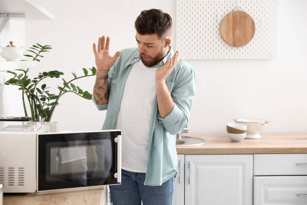 Shocked young man near microwave oven in kitchen - Фото, изображение
