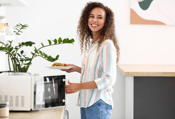 若いアフリカ系アメリカ人の女性は台所で電子レンジに食品とプレートを置く - 写真・画像
