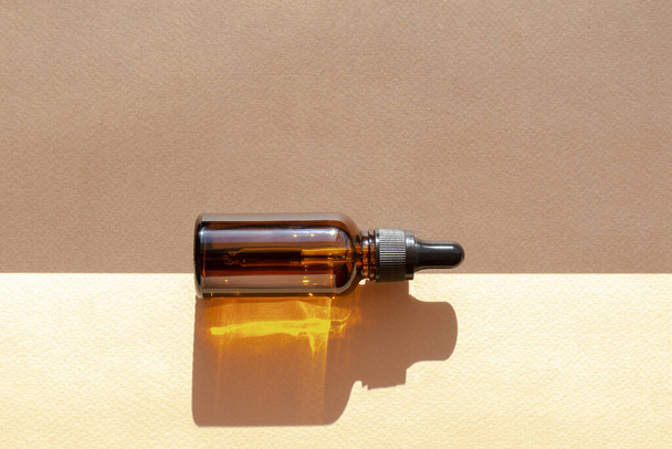 Mock-up of glass bottle with dropper lid, in rays of sunlight, lying at intersection of backgrounds of two colors: beige and brown. Rigid shadow of object falls on an abstract surface. - Fotó, kép