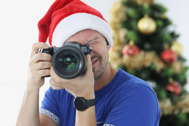 Kırmızı Noel Baba şapkalı bir adam Noel ağacının yanında fotoğraf çekiyor. - Fotoğraf, Görsel