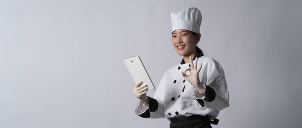 Asijská kuchařka drží smartphone nebo digitální tablet a obdržel objednávku z internetového obchodu nebo obchodní aplikace. usmívá se v kuchařské uniformě a stojí v bílém pozadí. Online obchodník s potravinami.  - Fotografie, Obrázek