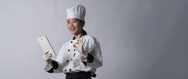 Mujer asiática chef sosteniendo teléfono inteligente o tableta digital y recibió el pedido de la tienda en línea o la aplicación comercial. sonriendo en uniforme de chef y de pie en fondo blanco. Comerciante de alimentos en línea.  - Foto, imagen