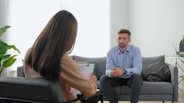 Mężczyzna pacjent z zaburzeniami psychicznymi siedzi na kanapie i rozmawia z terapeutą - Materiał filmowy, wideo