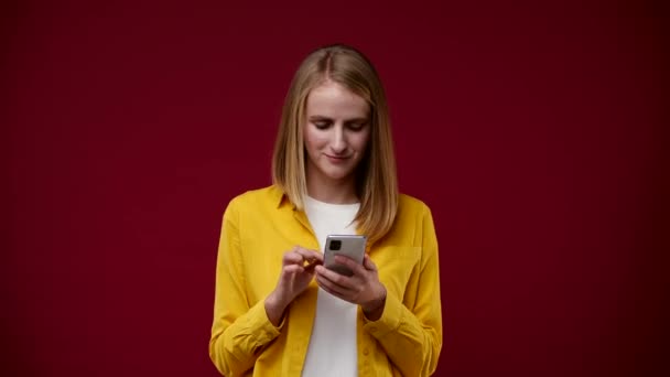 een meisje kijkt naar de telefoon en dan shows als en glimlacht, gekleed in een wit T-shirt en een geel shirt, de achtergrond is rood - Video