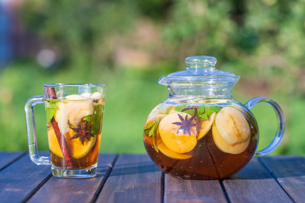 Healthy apple tea in a glass tea pot and mug in the summer garden on wooden table. Close up apple tea with cinnamon, cloves or mint leaves on nature background - Фото, зображення